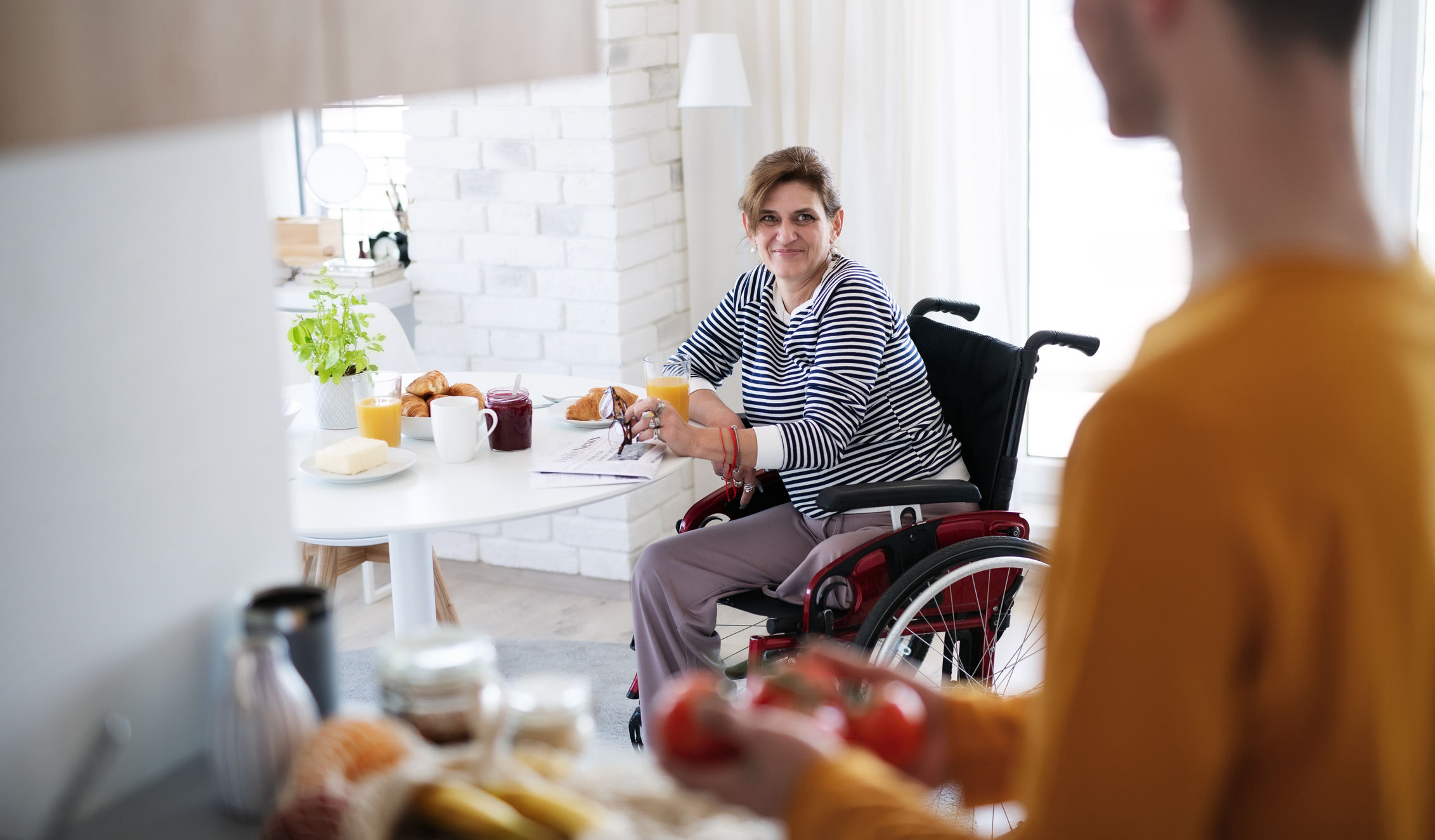 Disabled mature woman in wheelchair sitting at the table indoors at home, talking to friend.